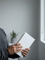 Person holding blank white book or magazine with empty cover for branding mockup in modern interior space with natural light and minimalist decor.