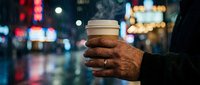 Mature man with dark skin holding warm coffee cup on rainy urban street at night with colorful city lights blurred in background, conveying comfort and solitude during cold weather.