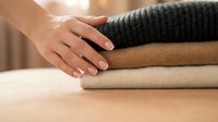 Hand touching stack of neatly folded knitted sweaters in neutral colors on wooden surface. Cozy autumn winter clothing organization and storage concept.