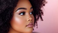 Close up portrait of young African American woman with natural curly hair and glowing makeup against pink background, side view beauty shot.