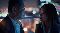 Professional mature businessman and young Asian businesswoman having intense conversation against blurred city lights background at night.