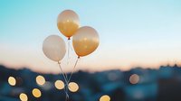 Pastel peach balloons floating against blurred sunset cityscape with bokeh lights creating dreamy atmospheric mood for celebration and special occasions.