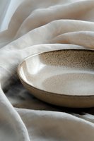 Handcrafted ceramic plate with speckled glaze on soft white linen fabric, creating minimalist composition with gentle folds and natural light for food styling.