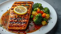 Grilled salmon fillet with lemon slice and glaze, served with colorful vegetable side of broccoli, carrots, and peas on white plate.