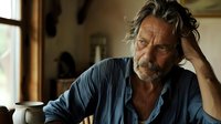 Mature man with gray hair and beard sitting pensively at wooden table near window, wearing blue casual shirt, natural morning light creates moody atmosphere.