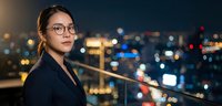 Professional Asian businesswoman with glasses standing on urban balcony at night with city lights bokeh background for corporate and career concepts.