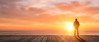 Man standing on wooden deck at sunset overlooking ocean horizon, contemplative pose during golden hour, suitable for motivational and lifestyle content.