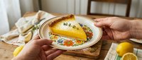 Hands holding vintage floral plate with slice of lemon tart garnished with thyme on rustic wooden table setting for homemade dessert presentation.