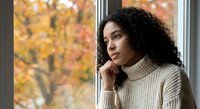 Young African American woman in cozy turtleneck sweater looking thoughtfully out window during autumn season with colorful fall foliage background.