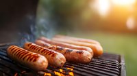 Juicy sausages grilling on barbecue with rising smoke and golden sunlight in background. Closeup of hot dogs cooking outdoors on summer evening.