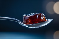 Red translucent jelly cube on silver spoon with bokeh background, macro food photography showing texture and light reflection for culinary and dessert designs.