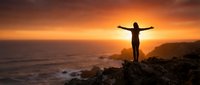 Woman silhouette standing on rocky cliff with arms outstretched against dramatic orange sunset over ocean, symbolizing freedom and triumph.