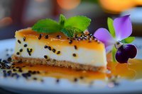 Creamy cheesecake slice garnished with caramel sauce, black sesame seeds, fresh mint leaves and edible purple flower on dark plate with bokeh background.