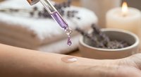 Lavender essential oil dropper dispensing purple oil onto hand with spa towels and candle creating relaxing aromatherapy wellness treatment atmosphere.