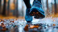 Running shoe splashing through puddle on wet autumn path with fallen orange leaves and water droplets in motion during rainy weather exercise.