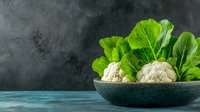 Fresh organic cauliflower heads with green leaves in gray ceramic bowl on turquoise table against dark textured background, healthy food concept.