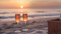 Two champagne glasses filled with sparkling wine on sandy beach at sunset with ocean waves and wicker basket creating romantic seaside celebration scene.