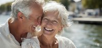 Happy senior couple laughing together outdoors by waterfront marina. Elderly man and woman with gray hair enjoying romantic moment in sunlight.