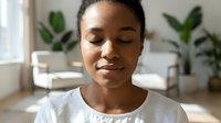 Young African woman with eyes closed in peaceful meditation pose, sitting indoors with potted plants in soft natural light, expressing calm and mindfulness for wellness and mental health content.