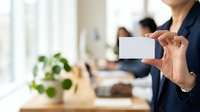 Woman in business attire holding blank white card in modern office workspace with colleagues and green plant, suitable for corporate branding and professional communications.