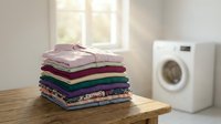 Clean folded laundry stack on wooden table in bright laundry room with white washing machine, domestic household chores and organization concept.