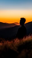 Silhouetted male figure standing in golden grassland at sunset, gazing toward distant mountains under warm orange sky. Contemplative outdoor portrait.