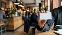 Businessman in dark suit holding blank white business card in modern office environment with blurred colleagues and warm lighting in background.