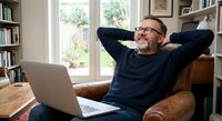 Relaxed mature man with beard and glasses sitting in leather chair with laptop, hands behind head in comfortable home office with bookshelves and natural light.
