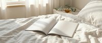 Open blank book mockup on a cozy white linen bed in a bright sunlit bedroom with soft curtains and a cup of tea in the background.