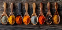 Colorful spices and seasonings in wooden spoons arranged in row on dark rustic wooden background, variety of ground turmeric, paprika, pepper and herbs.