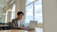 Young Asian businessman working on laptop in modern office space with large windows and natural light for professional business concepts.