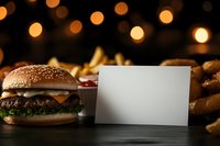 Blank white card mockup with juicy cheeseburger, french fries and blurred bokeh lights in dark restaurant setting, food menu presentation concept.