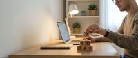 Mature man with light skin tone and facial hair, wearing a beige cardigan, stacking wooden blocks at a desk with laptop and notebook, representing business planning, risk management, and strategic growth in a modern home office setting.