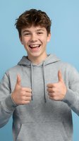 Happy teenage boy giving thumbs up gesture wearing gray hoodie on blue background expressing positive emotions and success.
