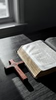 Open holy book with golden pages beside a wooden cross on a dark table, soft window light creating a spiritual, contemplative atmosphere.
