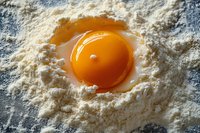 Fresh raw egg yolk in white flour nest on dark surface, macro food photography with soft natural lighting and textured details for cooking concept.