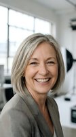 Mature woman with gray hair smiling at camera in bright office setting, wearing neutral blazer, suitable for corporate, business, and professional headshot applications.