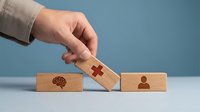 Close-up of hand placing wooden block with red cross symbol between brain and person icons on blue background, representing healthcare connection and medical services.