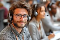 Smiling young Caucasian male customer service representative with headset and glasses in busy call center environment.