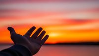 Hand reaching toward vibrant sunset sky over calm water, symbolizing hope, freedom and new beginnings in warm orange and pink twilight colors.