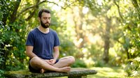 Young man with dark beard and hair, Caucasian ethnicity, wearing navy t-shirt, sitting cross-legged on log in forest meditating, surrounded by green trees and dappled sunlight for wellness and mindfulness content.