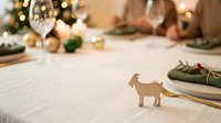 Wooden goat figurine on festive dining table with holiday decorations, garland, and bokeh lights for seasonal celebration styling.