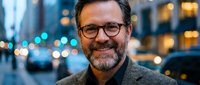 Smiling mature Caucasian businessman with glasses and beard standing on busy city street at dusk with bokeh lights for corporate headshot photography.