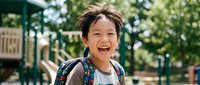 Happy Asian elementary school boy with backpack laughing joyfully at outdoor playground during sunny day, expressing pure childhood excitement and carefree moments.