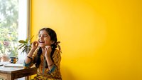 Young woman with dark hair and olive complexion sitting at desk by window against bright yellow wall, wearing mustard cardigan, smiling thoughtfully while resting chin on hands. Office or creative workspace setting.