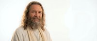 Smiling middle-aged Caucasian man with long curly hair and full beard wearing white robe on light background for spiritual or religious concepts.