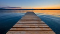 Wooden pier stretches into calm lake at sunset, reflecting orange sky and mountain silhouettes on tranquil blue water surface, creating serene landscape view.