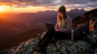 Woman working on laptop at mountain summit during golden hour sunset, remote work and digital nomad lifestyle concept for freelance professionals and outdoor adventure themes.