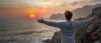 Man with arms outstretched standing on rocky cliff overlooking ocean at sunset, experiencing freedom and contemplation during golden hour with dramatic sky.