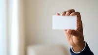 Male professional with dark skin tone holding blank white business card in right hand against neutral beige background, suitable for corporate branding and business identity projects.
